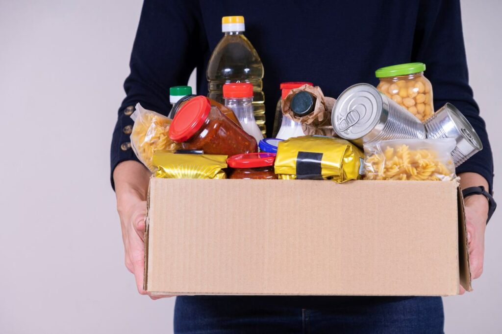 A variety of foods, including pasta noodles, sauce, and other packaged goods, are piled in a cardboard box carried by a person.
