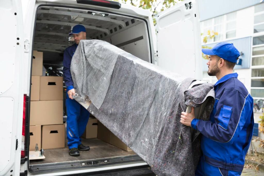 Two movers load a piece of wrapped furniture into the back of a van filled with other cardboard boxes. 