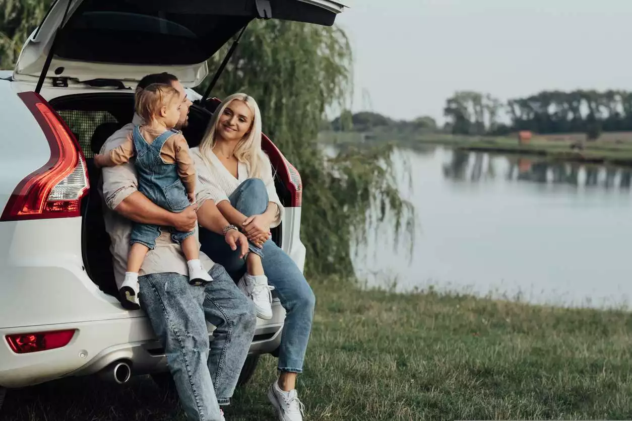 Young Parents with Little Daughter Sitting in Open Trunk of Car, Happy Family Enjoying Road Trip on Their SUV Car. 