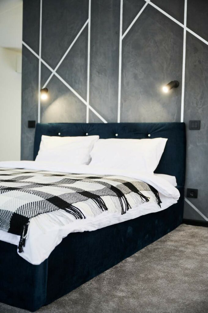 A black bed with white sheets and a checkered blanket in front of a gray accent wall with white geometric stripes