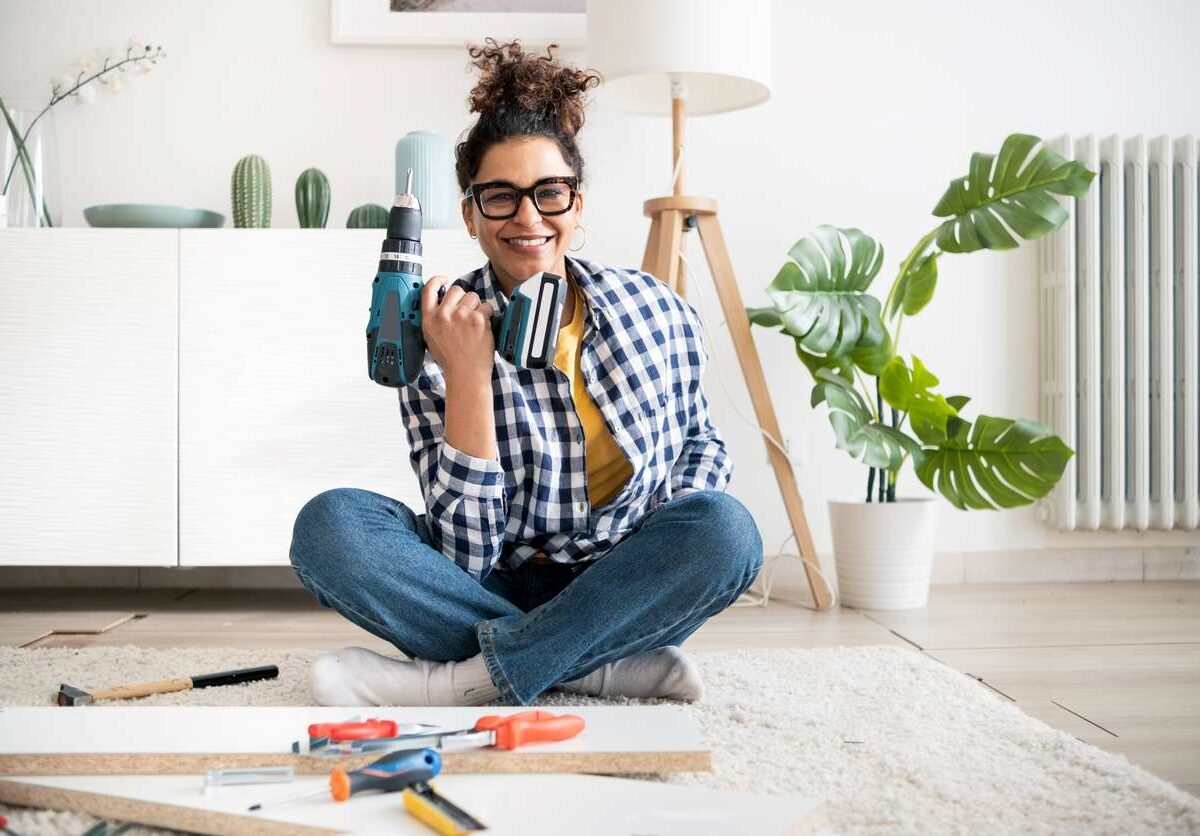 A woman in a checkered shirt smiles while holding a drill with other tools scattered in front of her on the floor