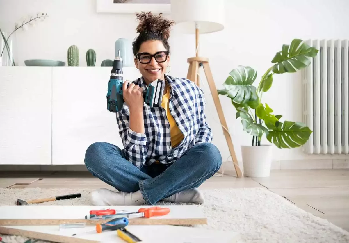 A woman in a checkered shirt smiles while holding a drill with other tools scattered in front of her on the floor