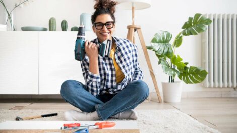 A woman in a checkered shirt smiles while holding a drill with other tools scattered in front of her on the floor