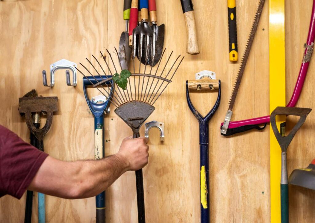 Homeowner reaches for rake from an organized garage wall with tools mounted on hooks.