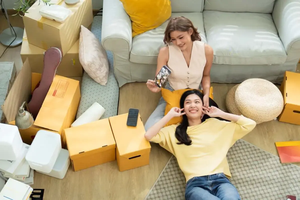 Two young women unpacking moving boxes in a cozy living room, smiling and relaxing among their items and furniture