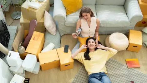 Two young women unpacking moving boxes in a cozy living room, smiling and relaxing among their items and furniture