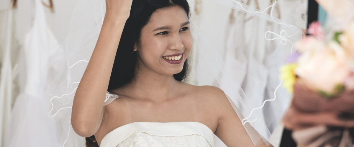 A smiling bride in a white wedding dress lifts her veil while standing in a bridal shop surrounded by hanging gowns.