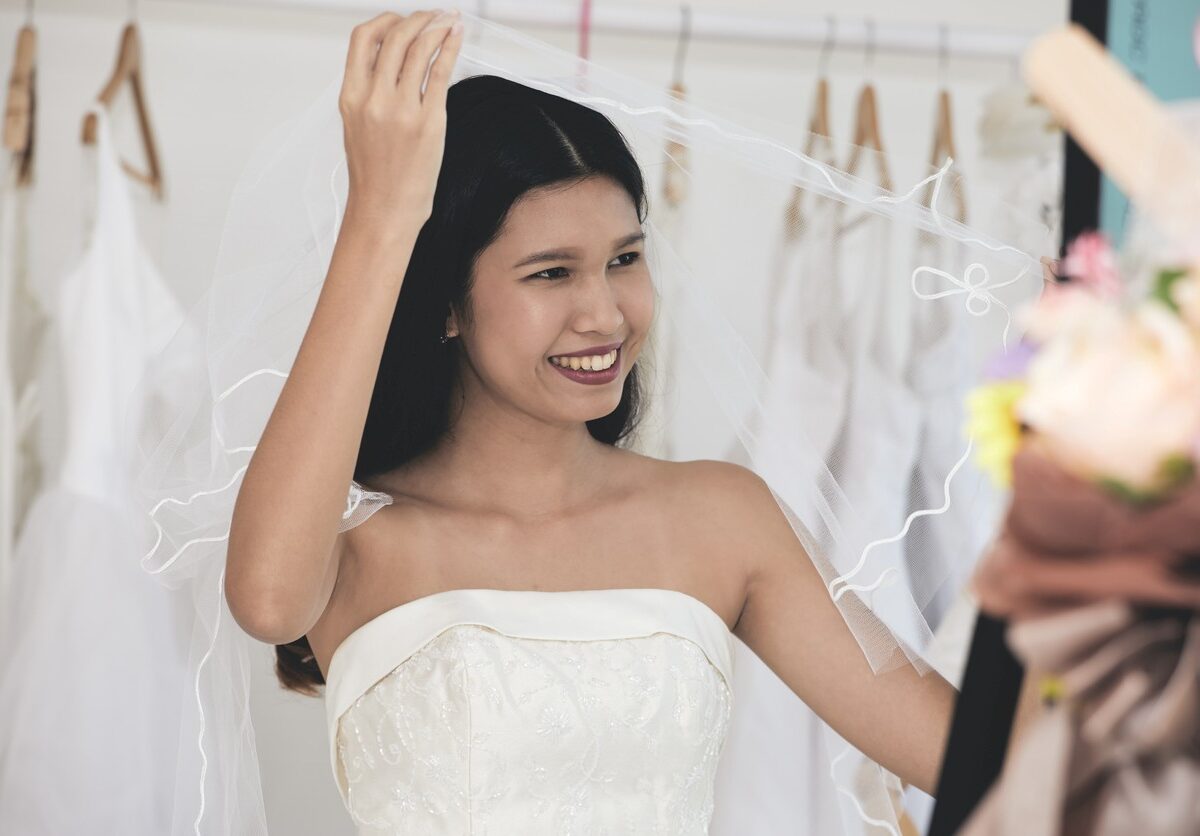 A smiling bride in a white wedding dress lifts her veil while standing in a bridal shop surrounded by hanging gowns.