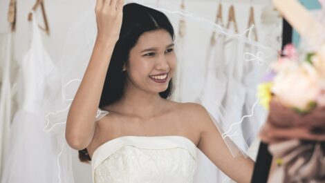 A smiling bride in a white wedding dress lifts her veil while standing in a bridal shop surrounded by hanging gowns.