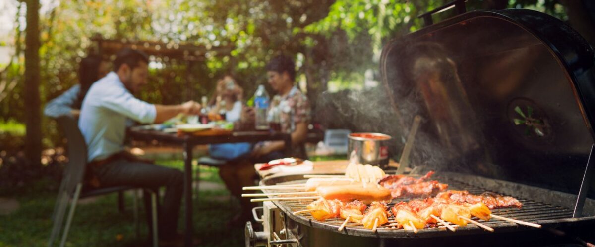 A family enjoying a backyard barbecue with grilled skewers cooking on a charcoal grill on a sunny day.
