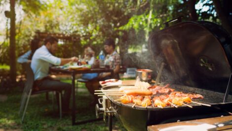 A family enjoying a backyard barbecue with grilled skewers cooking on a charcoal grill on a sunny day.