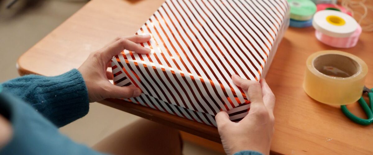 A person wrapping a gift with striped paper at a table, surrounded by ribbons, tape, and scissors.