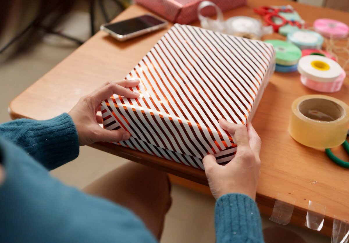 A person wrapping a gift with striped paper at a table, surrounded by ribbons, tape, and scissors.