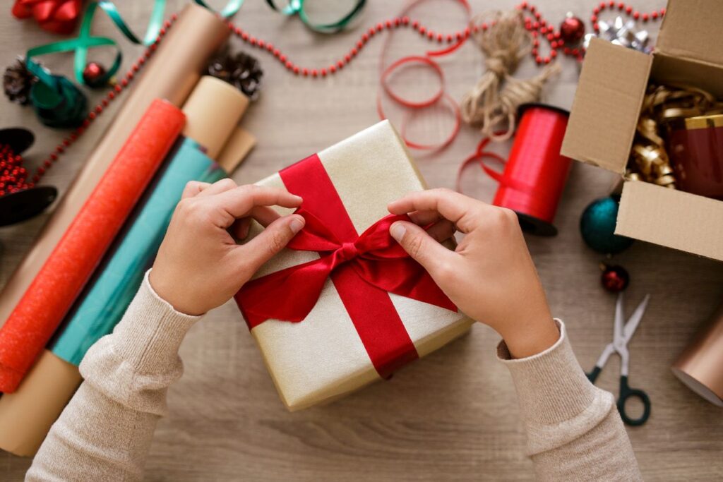 Hands tying a red ribbon on a wrapped gift surrounded by wrapping paper, scissors, ribbons, and holiday decorations.
