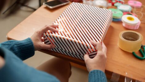 A person wrapping a gift with striped paper at a table, surrounded by ribbons, tape, and scissors.