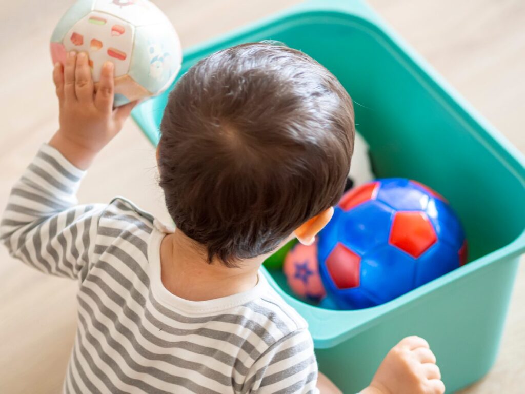 A toddler places soft toy balls into a green storage bin during cleanup time in a bright, tidy room.
