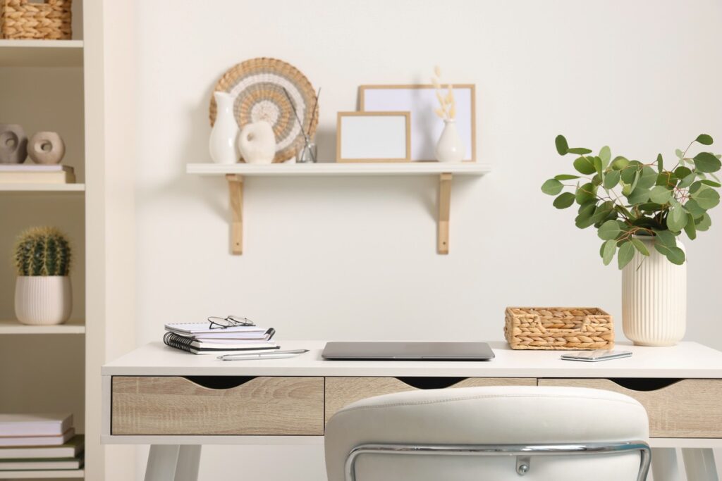 A modern home office with a white desk, laptop, notebooks, and a plant in a vase, with shelves and minimalist decor.