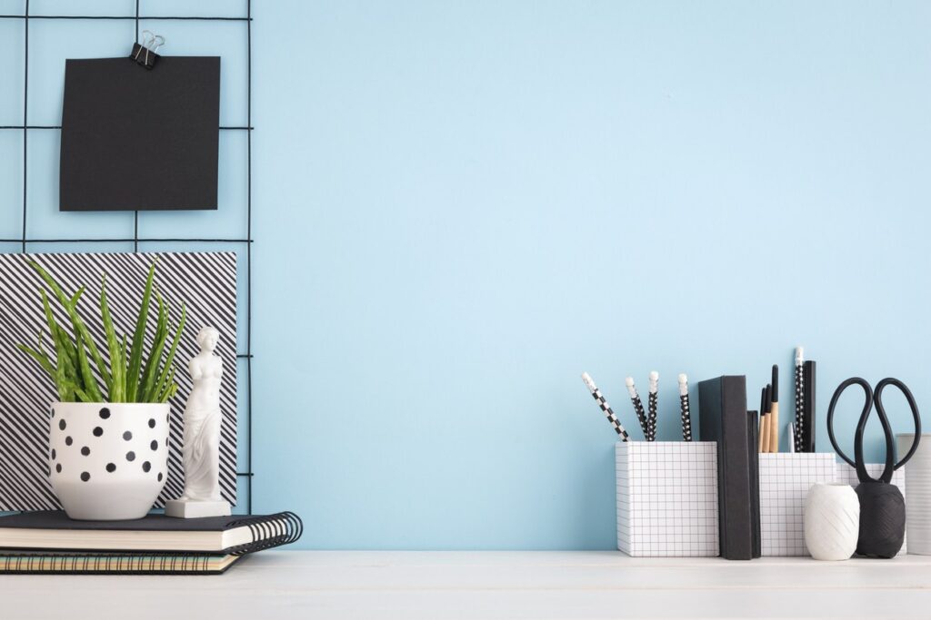 A minimalist desk with a plant in a polka dot pot, notebooks, pencil holders, and scissors against a light blue wall.