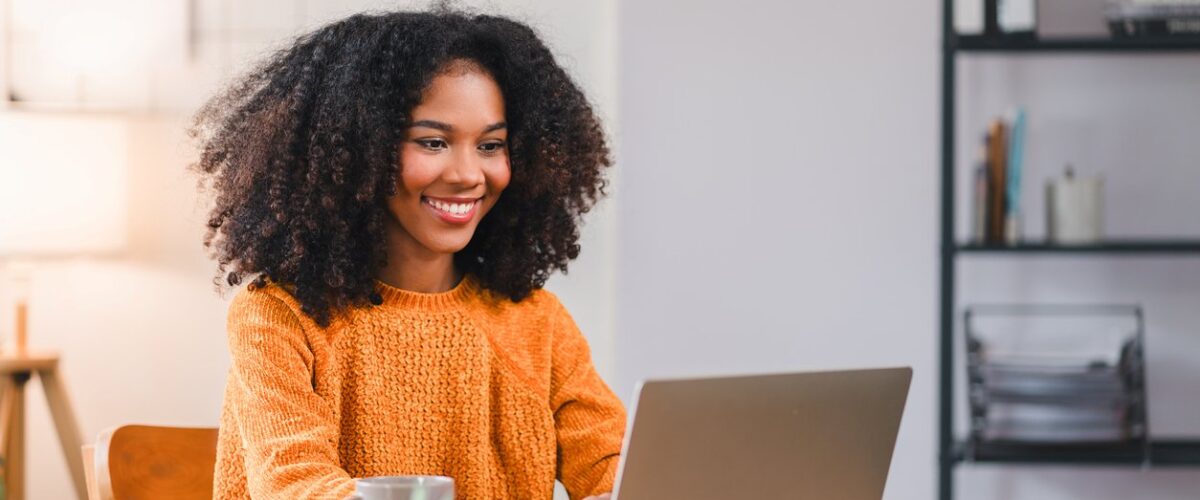 A smiling woman in an orange sweater works on a laptop at a wooden desk with books and a plant.