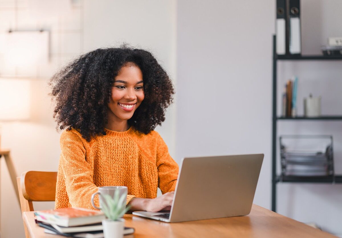 A smiling woman in an orange sweater works on a laptop at a wooden desk with books and a plant.