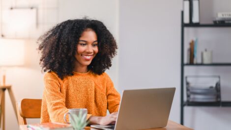 A smiling woman in an orange sweater works on a laptop at a wooden desk with books and a plant.