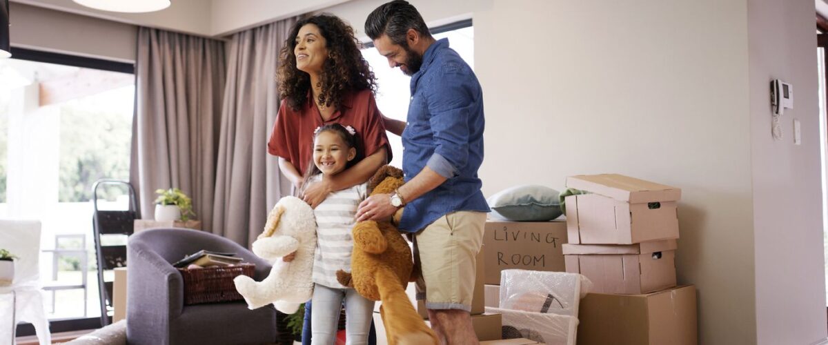 A happy family carrying stuffed animals while moving into a new home, surrounded by boxes and living room furniture.