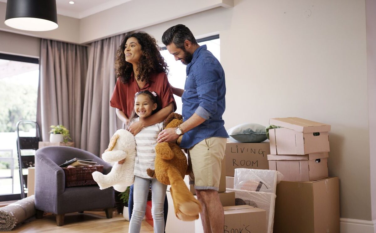 A happy family carrying stuffed animals while moving into a new home, surrounded by boxes and living room furniture.