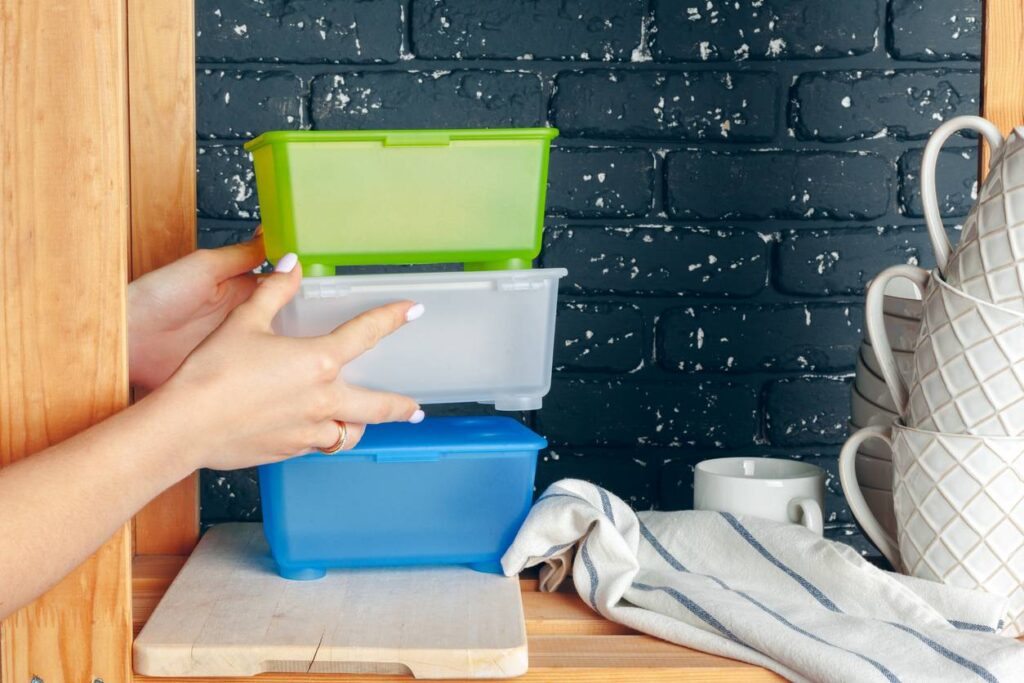 Hands stacking colorful plastic storage containers on a wooden shelf in a kitchen with mugs and a striped towel nearby.