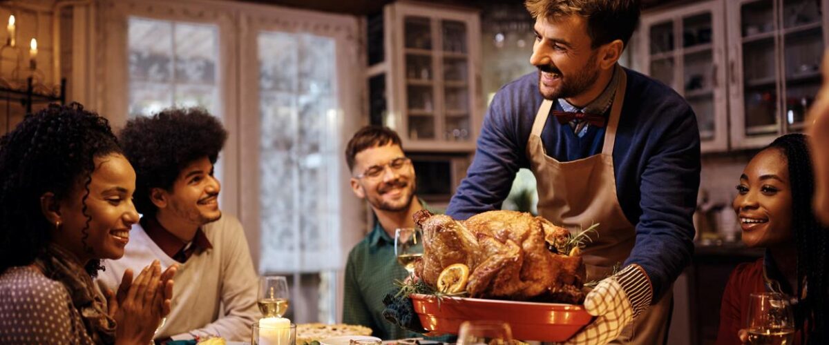 A smiling group of friends sits around a decorated Thanksgiving table as a man proudly brings out a roast turkey.