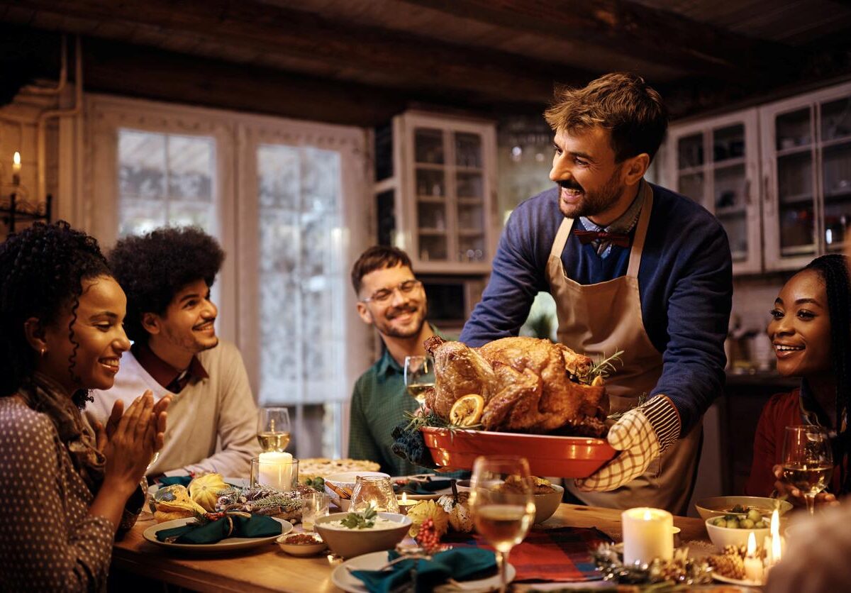 A smiling group of friends sits around a decorated Thanksgiving table as a man proudly brings out a roast turkey.