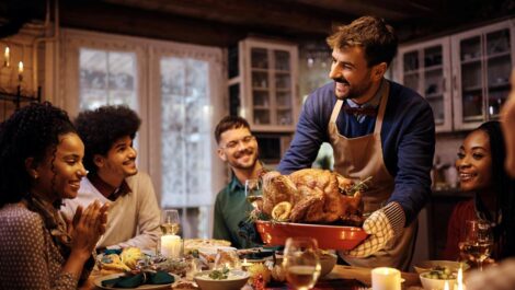 A smiling group of friends sits around a decorated Thanksgiving table as a man proudly brings out a roast turkey.