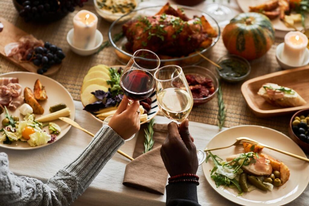 Two people clink glasses of red and white wine over a festive Thanksgiving table filled with food and fall decor.