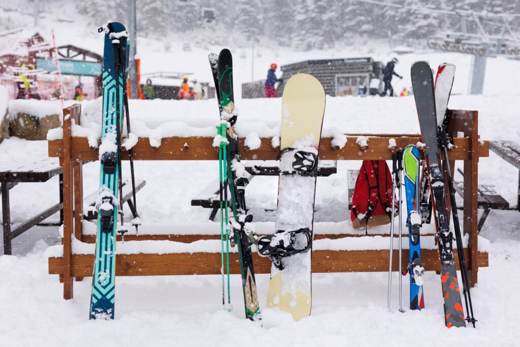 Skis and a snowboard standing in a snowy rack at a ski resort, covered in fresh snow with people and a lift.