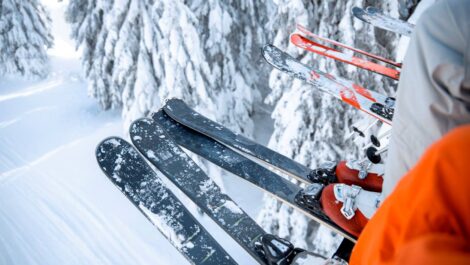 The legs and feet of a group of people on a ski lift above a snowy forest with their skis covered in snow.