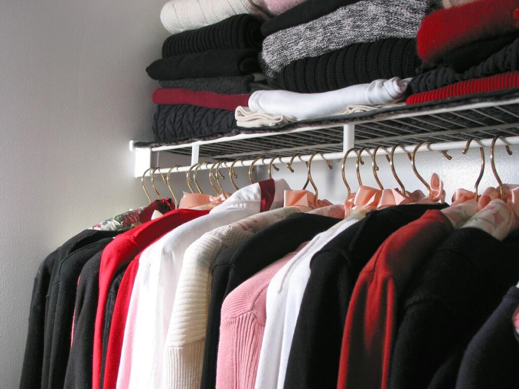 Neatly organized sweaters and winter tops hanging on gold hangers with folded clothes stacked on a shelf in a tidy closet. 